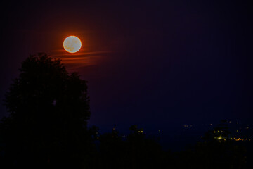 Obraz premium Bright full moon rises over dark landscape with distant lights in the evening sky