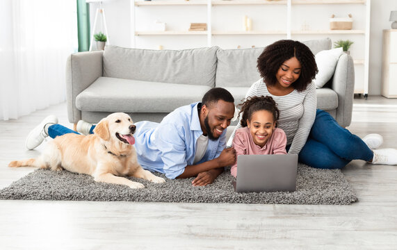 Portrait of happy loving black family lying on floor carpet with labrador, using laptop and watching video or movie, browsing internet, spending time together at home in living room