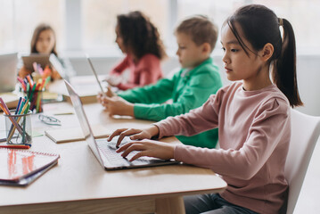 Korean Little Girl Using Laptop Sitting At Desk With Multiracial Classmates In Modern Classroom Indoor. E-Learning Website And Modern Internet Education Service. Selective Focus