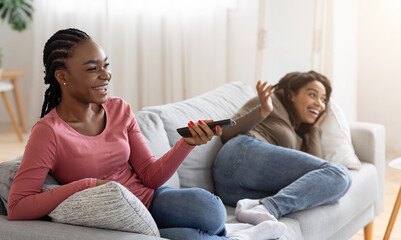Obraz premium Two cute black ladies having fun on weekend at home, watching TV on sofa at living room, empty space. Happy african american women friends laying on couch, watching funny content on television