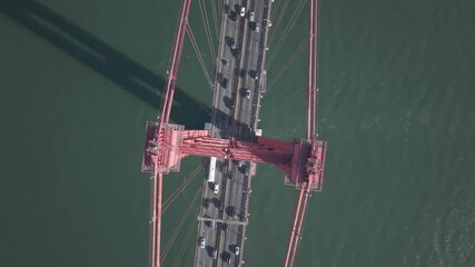 Stunning top-down aerial view of the iconic 25 de Abril Bridge in Lisbon, Portugal, showcasing the bridge's red steel structure, urban traffic, and deep green waters of the Tagus River. Perfect for th