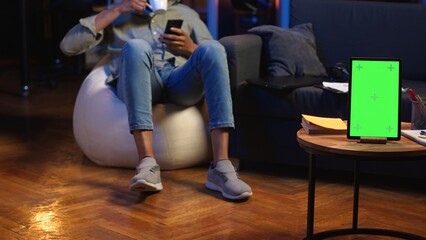 Black young person browses laptop and phone next to green screen, feeling casual and relaxed on the...