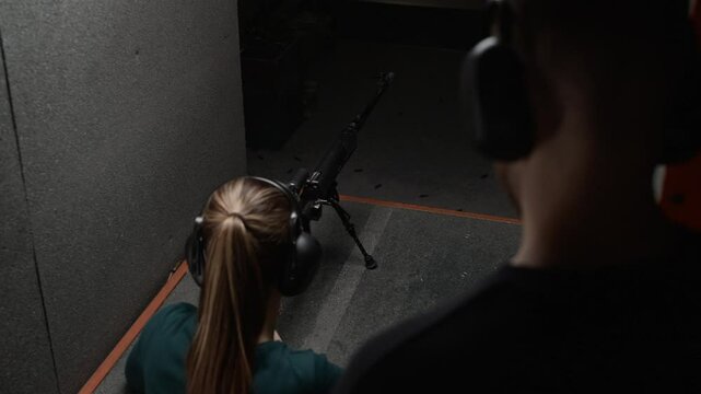 Over the shoulder view of male instructor wearing earmuffs guiding female trainee shooting rifle in range