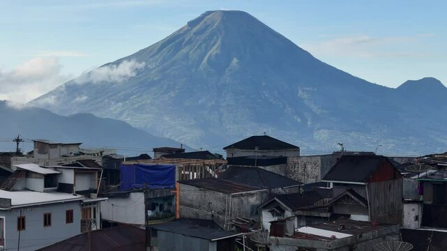 Mount Sindoro rises majestically in the background as rooftops of a densely-packed village in Central Java, Indonesia, fill the foreground. The morning light enhances the contrast between the rugged m