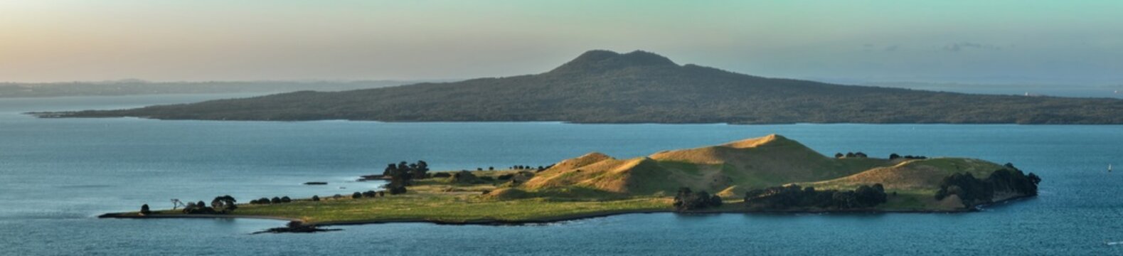 Panoramic view of Browns Island and Rangitoto and sea in New Zealand. The foreground island's hills are bathed in warm, golden light.