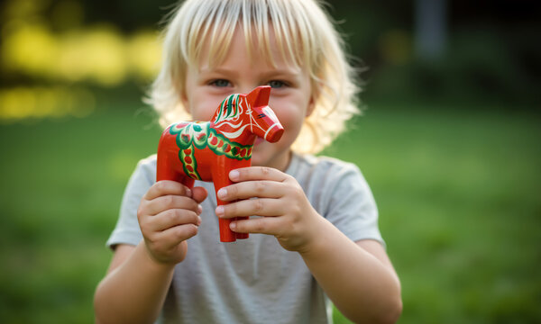 Young blonde child holding a red Dala horse toy outdoors. Portrait with Swedish folk art symbol. Swedish Dalecarlian toy horse souvenir. Symbol of Sweden.
