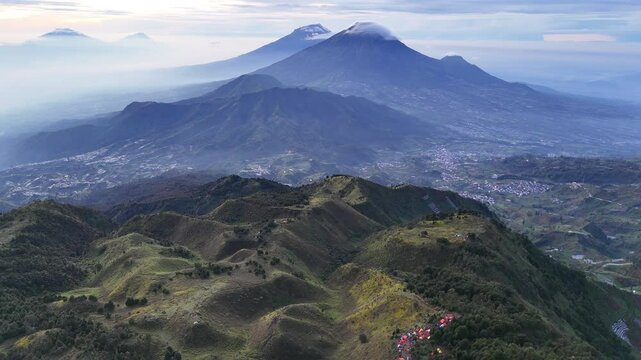 A stunning aerial view of a mountain range in Central Java, Indonesia, featuring Mount Sindoro and Mount Sumbing as the prominent peaks. The landscape showcases a mix of lush green hills, agricultural