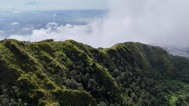 A breathtaking view of Mount Ungaran's lush green ridgeline in Central Java, Indonesia. The steep mountain slopes are covered in rich vegetation, partially cloaked by drifting clouds and mist.