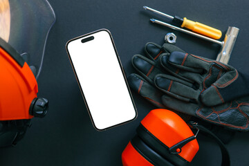 Smartphone with blank screen surrounded by construction tools, gloves, helmet, and ear protection on black background. Industrial safety concept