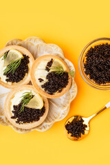 Plate of tasty tartlets with black caviar on yellow background