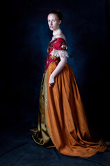 A red-haired Renaissance woman wearing an embroidered silk ensemble against a studio backdrop