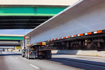 Obraz premium Truck carries long concrete slabs along highway surrounded by overpasses on clear day.