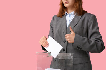 Voting young woman showing thumb-up near ballot box on pink background