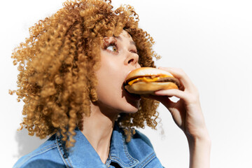 Young woman with curly hair excitedly holding a hamburger, dressed in a denim jacket against a white background, symbolizing indulgence and fast food culture