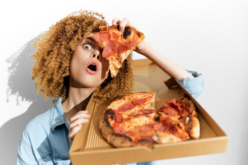 Surprised young woman with curly hair holding a slice of pizza, showcasing excitement and enjoyment for delicious food