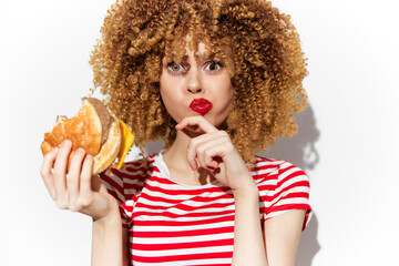 Young woman with curly hair in stripes holding a burger, playfully shushing with a surprised expression, ideal for healthy eating and fast food concepts