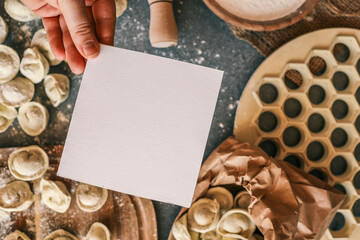 Blank white card held above a rustic kitchen table with homemade dumplings, flour, and cooking utensils, perfect for recipe notes or invitations
