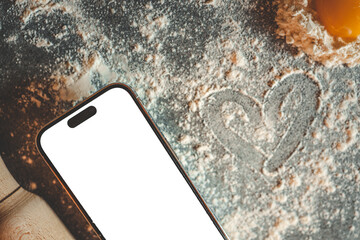 Top view of raw dumplings with flour, egg yolk, heart shape drawn in flour, rolling pin, and smartphone with blank screen on a kitchen table