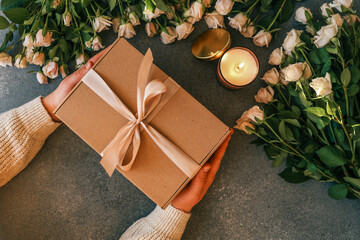 Hands holding a gift box with a satin ribbon, surrounded by white roses, a burning candle, and soft cozy fabric on a dark textured background