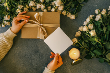 Hands holding a gift box and blank card surrounded by cream roses and a lit candle. Cozy flatlay with warm light and romantic mood, perfect for celebration or message