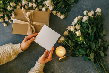 Hands holding a blank greeting card above a gift box with satin ribbon, surrounded by beige roses and a glowing candle on a dark stone background