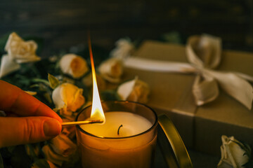 Close-up of a match lighting a candle, surrounded by soft beige roses and a gift box with ribbon in a warm cozy atmosphere