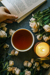 Cozy flat lay with a cup of tea, open book, bouquet of pale roses, a lit candle, and a gift box with ribbon on a stone surface