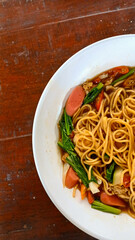 Top view of a white bowl filled with stir-fried noodles, sausage slices, leafy greens, cabbage, and chili in a savory sauce, placed on a rustic wooden table.