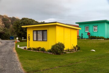 Colorful holiday cabins line a gravel road in Awakino, Waikato, New Zealand. Tourists stay in the rental units for vacation. The cabins are painted yellow, orange, green, pink, and blue.
