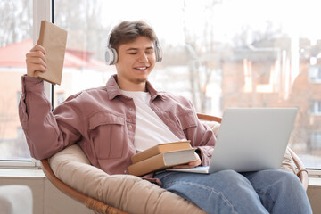 Male student with headphones and books studying online in armchair at home