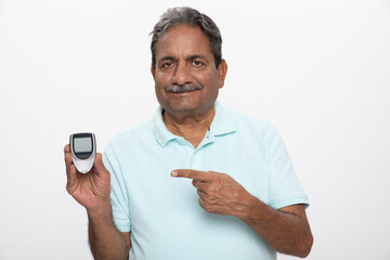 Blood sugar machine. Portraits of indian senior man showing blood glucose meter while standing grey background