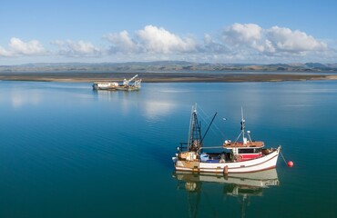 Fototapeta premium Fishing boats rest in calm waters near the in the Kawhia Harbour, Kawhia, Waikato, New Zealand. Fishing boat moored, while a dredge boat is anchored further out.