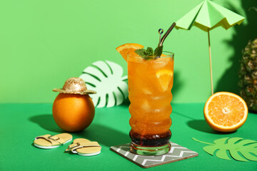 Glass of tasty cocktail with orange, umbrella, paper palm leaves and flip flops on green background