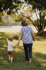 Fototapeta premium Grandmother Walking Hand in Hand with Young Grandson in Sunny Park, Outdoor Family Bonding Moment Captured from Behind