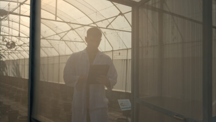 A scientist in a lab coat uses a tablet inside a greenhouse