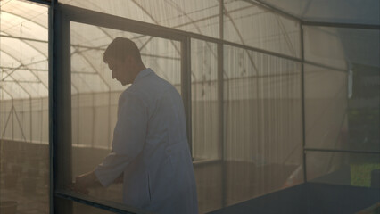 A scientist in a lab coat uses a tablet inside a greenhouse