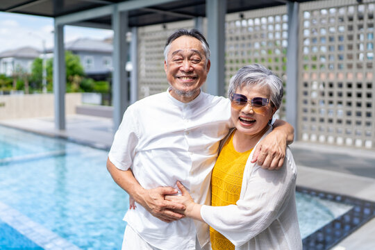 Portrait of happy grey-haired senior couple standing by swimming pool while travelling together, asian elderly people live wellbeing life enjoy summer vacation joyful retirement healthy lifestyle