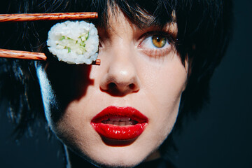 Portrait of a young woman with short black hair, holding sushi with chopsticks, emphasizing bold red lips and a striking expression.