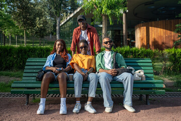 Portrait of four happy black best friends relaxing and hugging on park bench, enjoying of meeting, look at camera. Close friendship, share joyful moments together on stroll, rest and relax after work