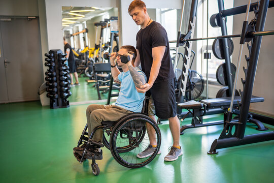 Trainer assists young man in wheelchair on personal session at gym, exercise technique monitoring moves. Disability support, rehabilitation spinal injury recovery, adaptive fitness, physical therapy