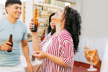 Young latin woman with curls, dancing with friends on a terrace, holding a bottle of beer