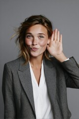 A woman in a blazer playfully raising her hand to her ear, inviting a conversation.
