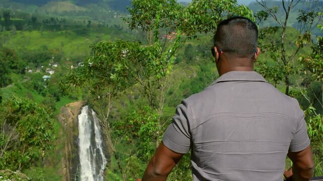 Tourist wearing a gray polo shirt, standing behind a railing and admiring Ramboda Falls, surrounded by lush green tea plantations in Sri Lanka