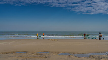 Obraz premium A group of people is pulling fishing nets out of the ocean. Turquoise waves spread across the sand. Reflection. Low tide. Puddles of water in the foreground. Blue sky, clouds. Copy space. Madagascar. 