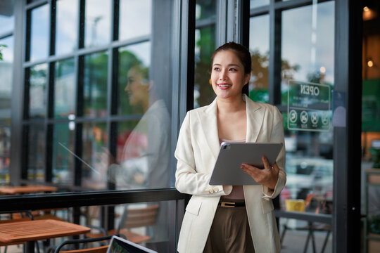 Happy entrepreneur holding a digital tablet, smiling while looking outside a cafe with an open sign, enjoying the vibrant urban atmosphere