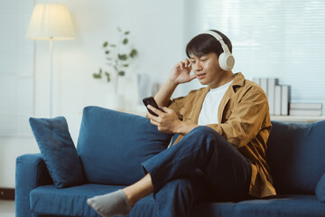 Obraz premium Relaxed young man enjoying music through wireless headphones while selecting songs on his smartphone, sitting comfortably on a sofa in his modern living room