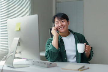 Young Asian man working remotely at a desk, using a desktop computer, chatting on the phone, and...