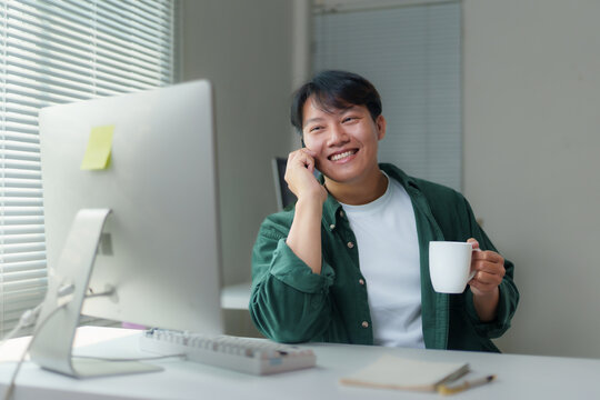 Young Asian man working remotely at a desk, using a desktop computer, chatting on the phone, and enjoying a cup of coffee