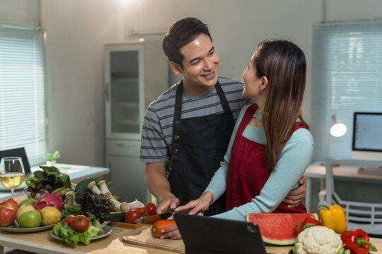 Happy asian couple wearing aprons preparing healthy meal with fresh vegetables together in kitchen