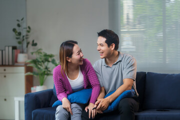 Obraz premium Happy Asian couple laughing and enjoying each other's company while sitting together on a cozy sofa in their inviting living room